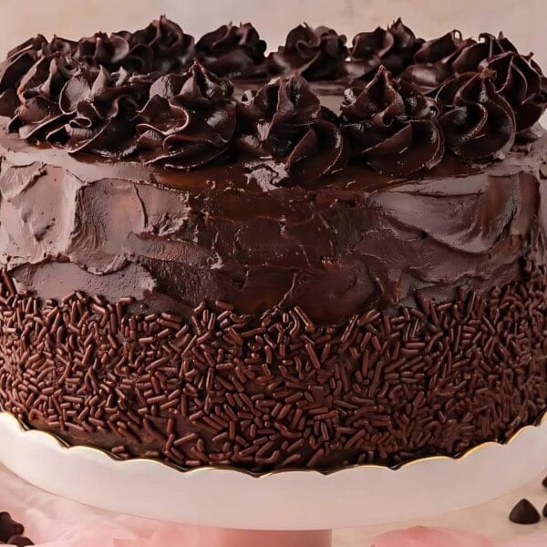 A chocolate cake with dark frosting and chocolate sprinkles, topped with decorative swirls, is placed on a white cake stand. Surrounding it are plates and pieces of chocolate.