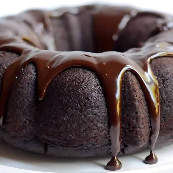 A chocolate bundt cake with glossy chocolate glaze drips down the sides, on a white plate.