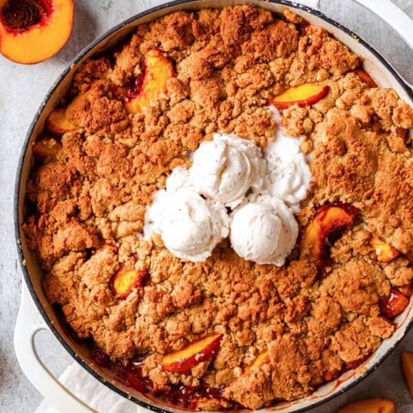 A white dish contains a peach cobbler topped with three scoops of vanilla ice cream, surrounded by fresh peaches and white flowers.