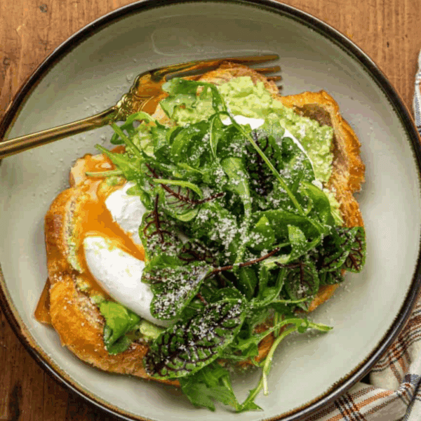 A plate with avocado toast topped with a poached egg and mixed greens, served with a fork on a wooden table next to a plaid napkin.
