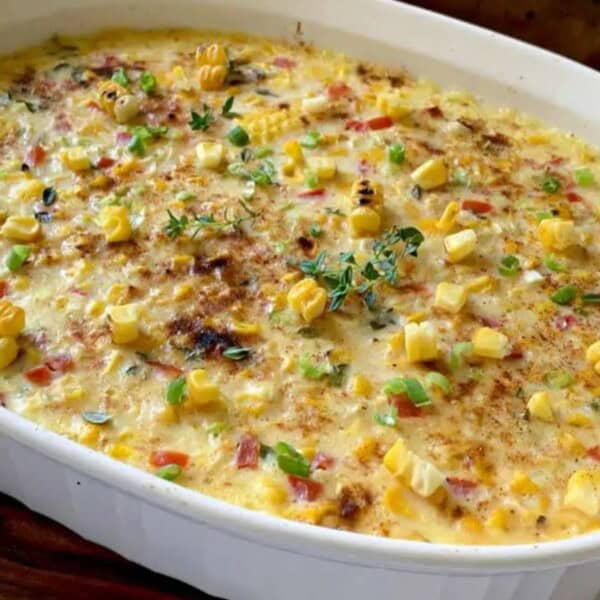 A baked casserole with a creamy corn filling, topped with diced red and green vegetables and fresh herbs, served in a white baking dish on a wooden surface.