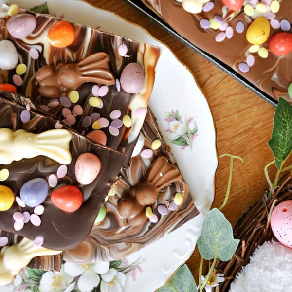 The image displays a chocolate bark topped with colorful candy eggs, sprinkles, and bunny-shaped chocolates. It is set on floral plates on a wooden table, surrounded by decorative eggs and vibrant flowers.