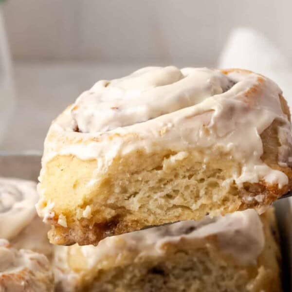 A frosted cinnamon roll is lifted with a spatula from a baking tray, and additional rolls are visible in the background.