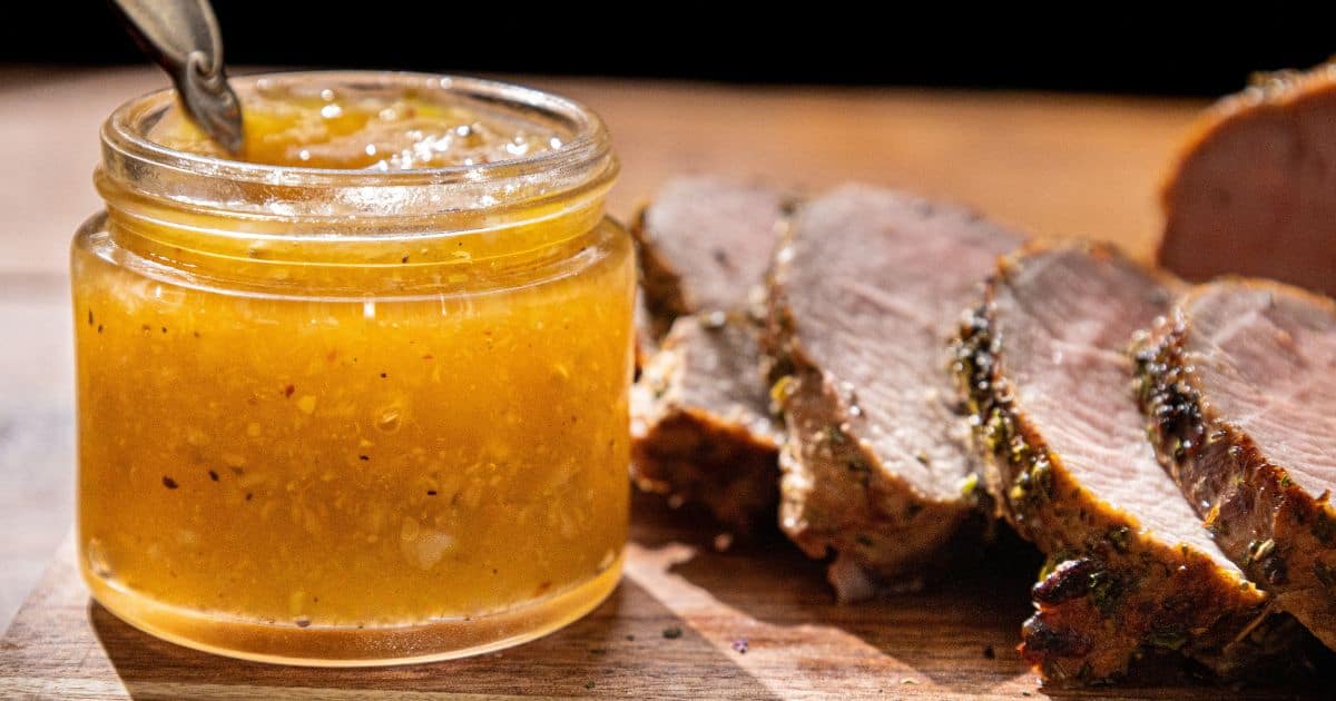A jar of yellow chutney with a spoon inside sits on a wooden surface next to several slices of cooked, seasoned meat.