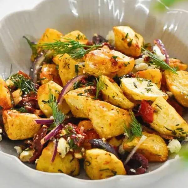 A creamy bowl of roasted potatoes mixed with red onions, olives, herbs, and feta cheese, garnished with fresh dill.