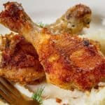 The image shows two chicken drumsticks on a bed of mashed potatoes, garnished with dill, alongside a fork on the plate.