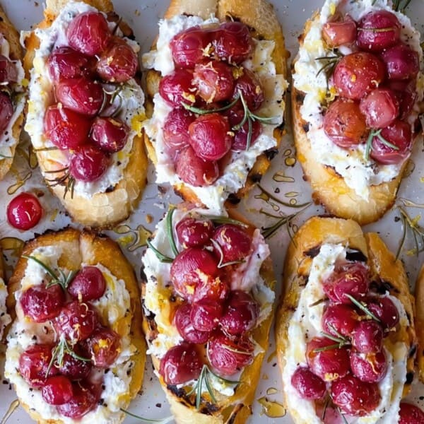 Crostini topped with roasted red grapes, whipped ricotta, rosemary, and honey drizzle. The toasted slices of bread are neatly arranged, highlighting the vibrant red grapes and creamy ricotta on a white surface.