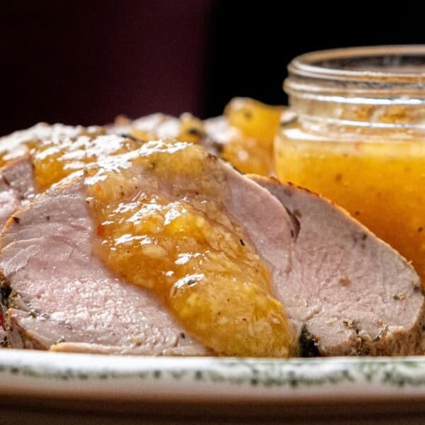 Sliced pork tenderloin topped with a golden sauce is arranged on a plate beside a small jar of sauce with a spoon.