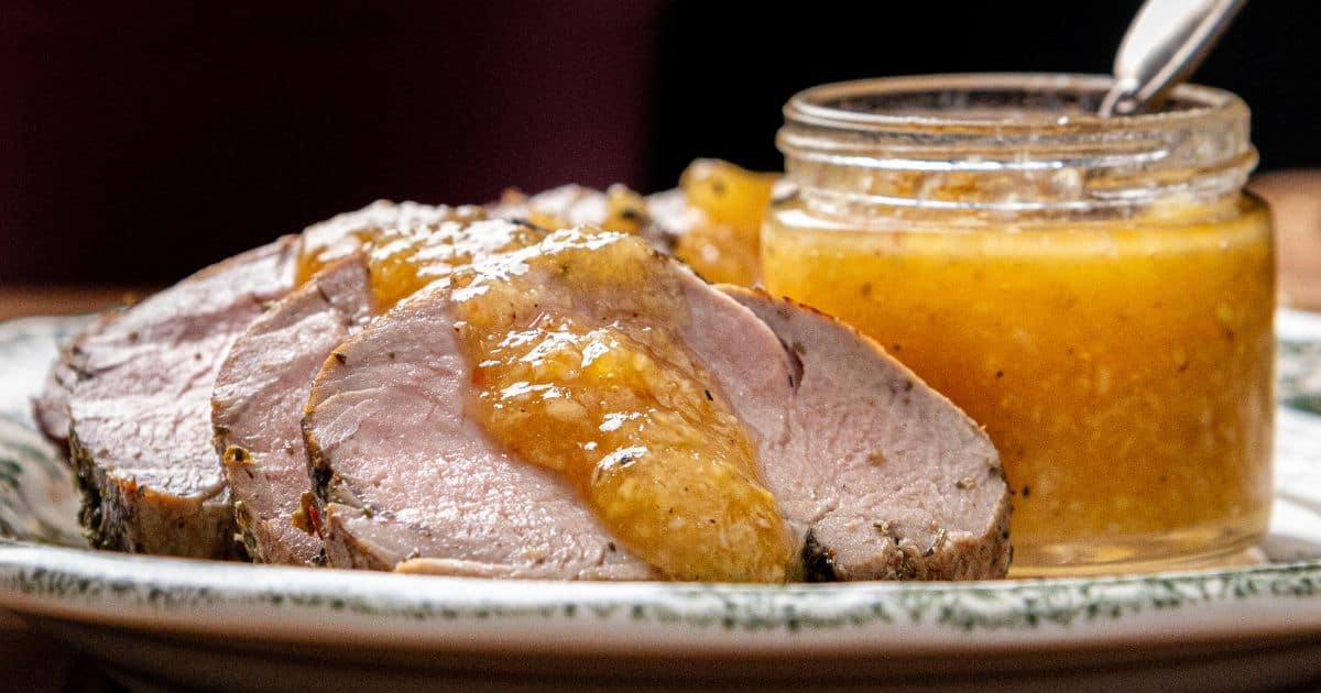 Sliced pork tenderloin topped with a golden sauce is arranged on a plate beside a small jar of sauce with a spoon.