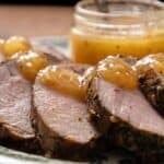 A plate with slices of pork tenderloin topped with sauce, accompanied by a jar of sauce in the background.