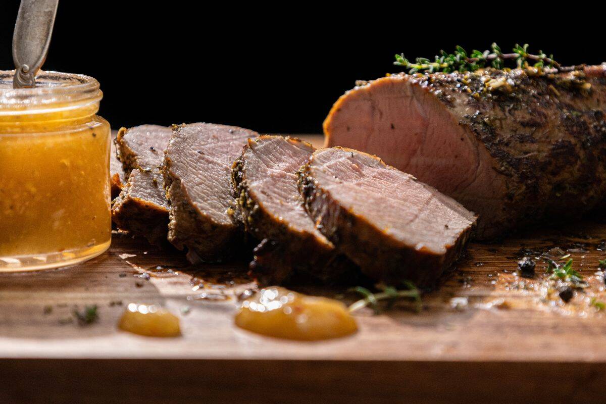 Sliced roast pork tenderloin arranged on a wooden board with sprigs of fresh herbs, beside a jar of yellow sauce or jelly.