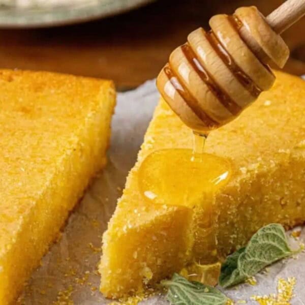 A honey dipper drizzles honey onto a slice of cornbread, with two additional slices and several mint leaves arranged on parchment paper.
