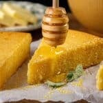 Slices of cornbread rest on parchment paper, with honey being drizzled over them. A plate with butter sits in the background.