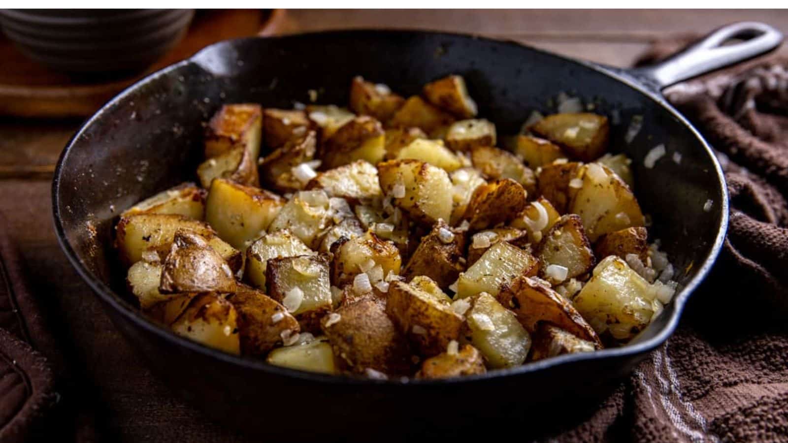 Golden-brown roasted potatoes with crispy edges, sprinkled with minced onions, served in a cast iron skillet on a brown cloth backdrop.