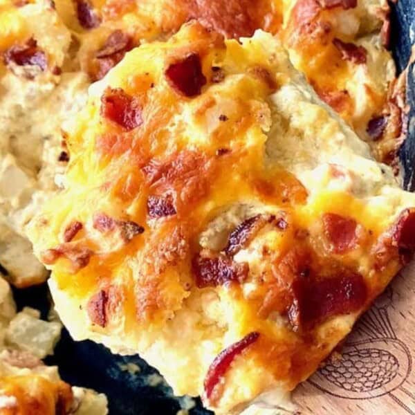 The image features a close-up of a cheesy baked dish with bacon pieces in a skillet. A wooden spatula is lifting a portion, displaying the melted cheese inside.