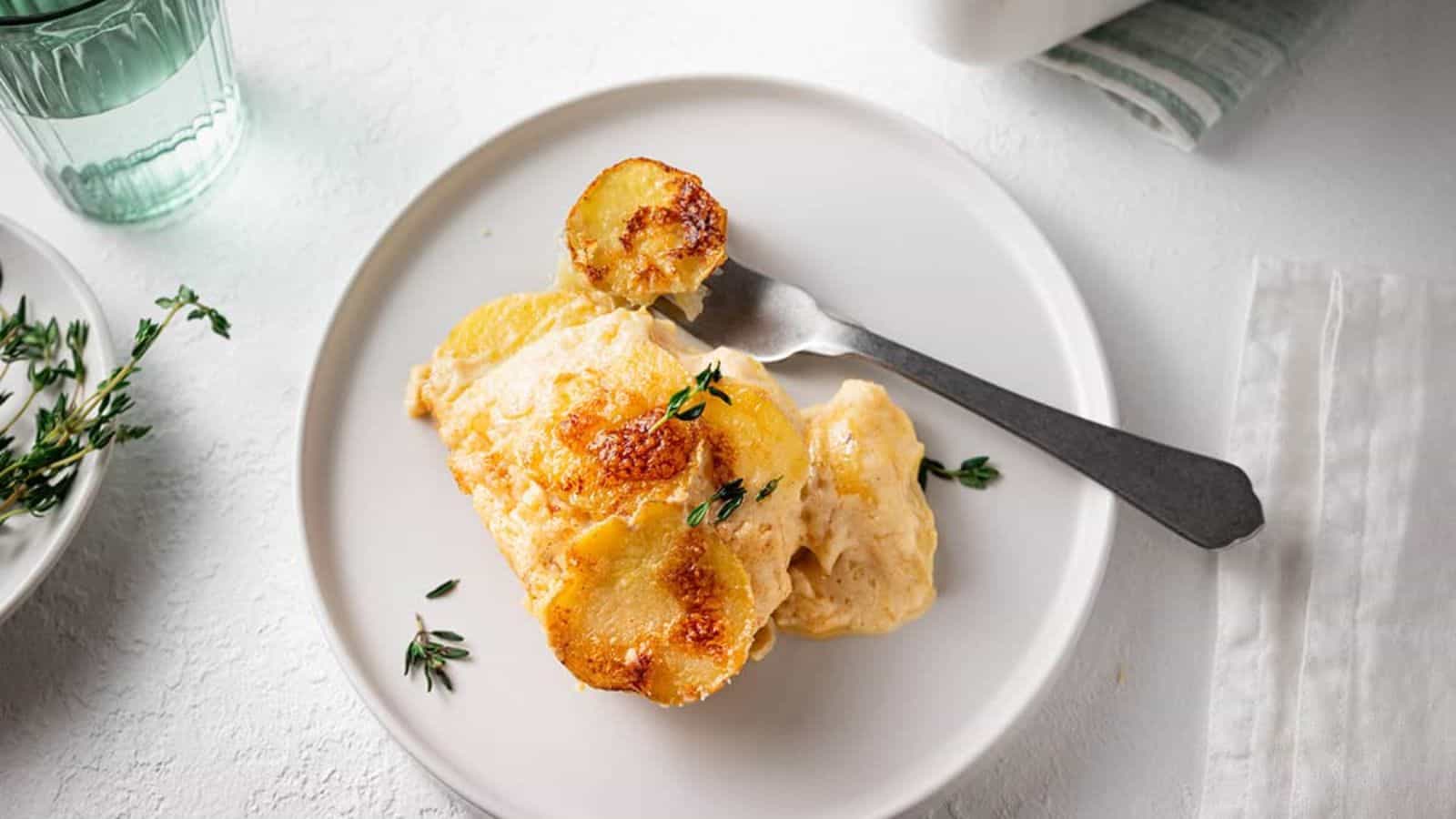 A creamy potato dish garnished with thyme, served on a white plate with a fork, accompanied by a glass of water and a side of herbs.