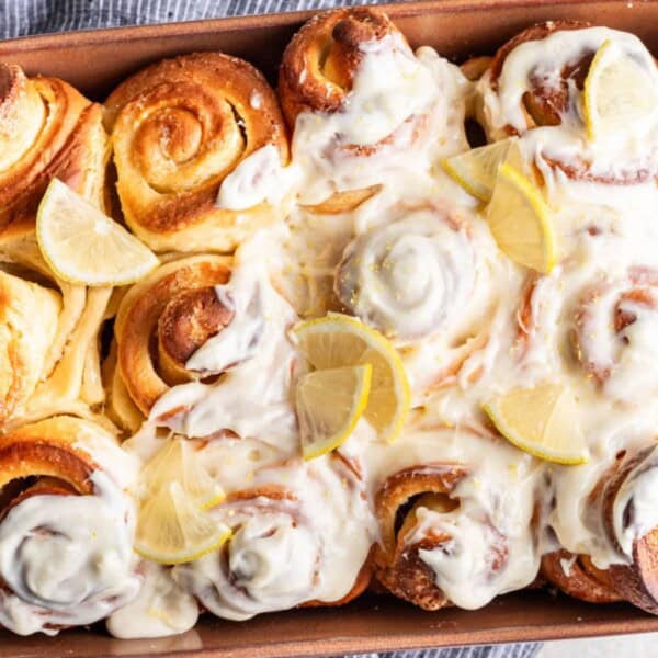 A baking dish filled with frosted cinnamon rolls is garnished with lemon slices and placed on a textured surface. The background includes plates and halved lemons.