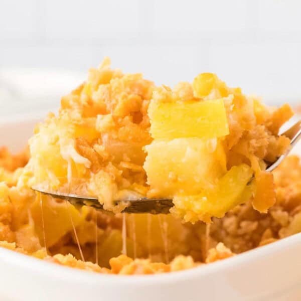 A serving spoon lifting a portion of casserole with visible pineapple chunks, cheese, and crumb topping from a white baking dish.