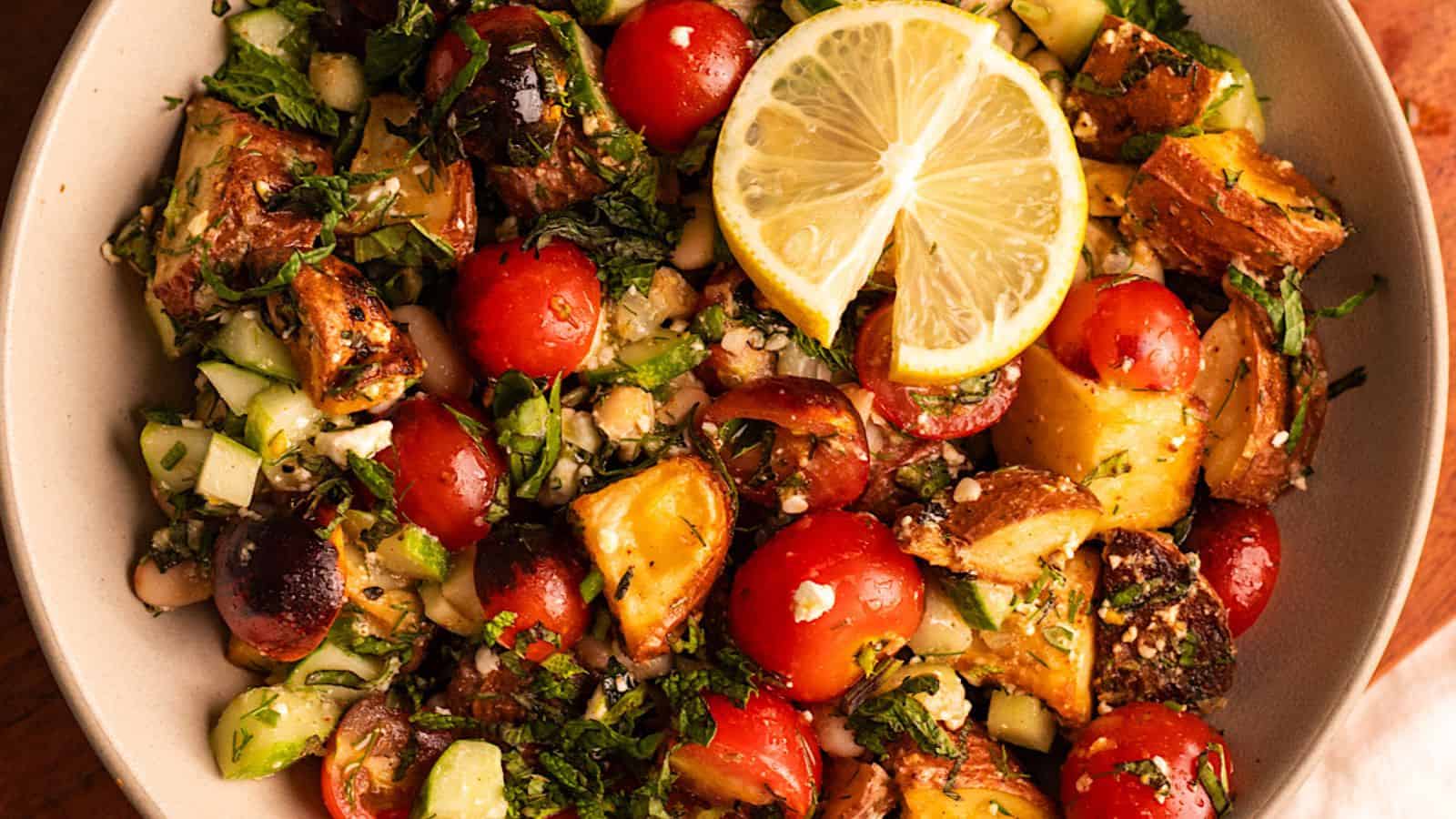 A vibrant salad with cherry tomatoes, roasted potatoes, cucumbers, herbs, and a lemon slice on top, served in a beige bowl.