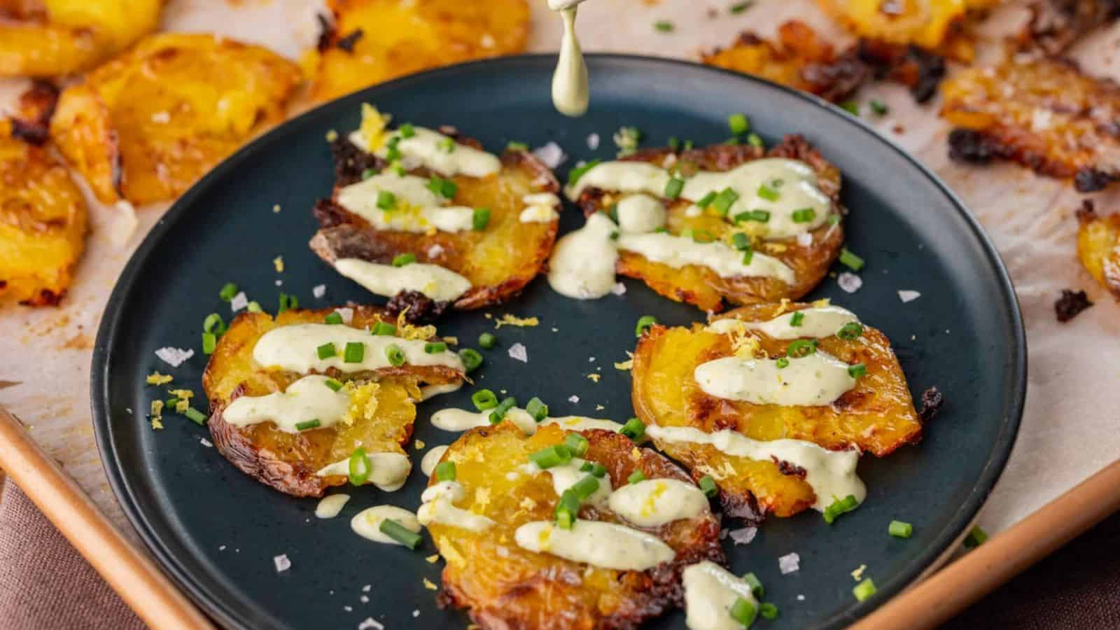 A black plate holds golden, crispy smashed potatoes drizzled with creamy sauce and garnished with green onions.