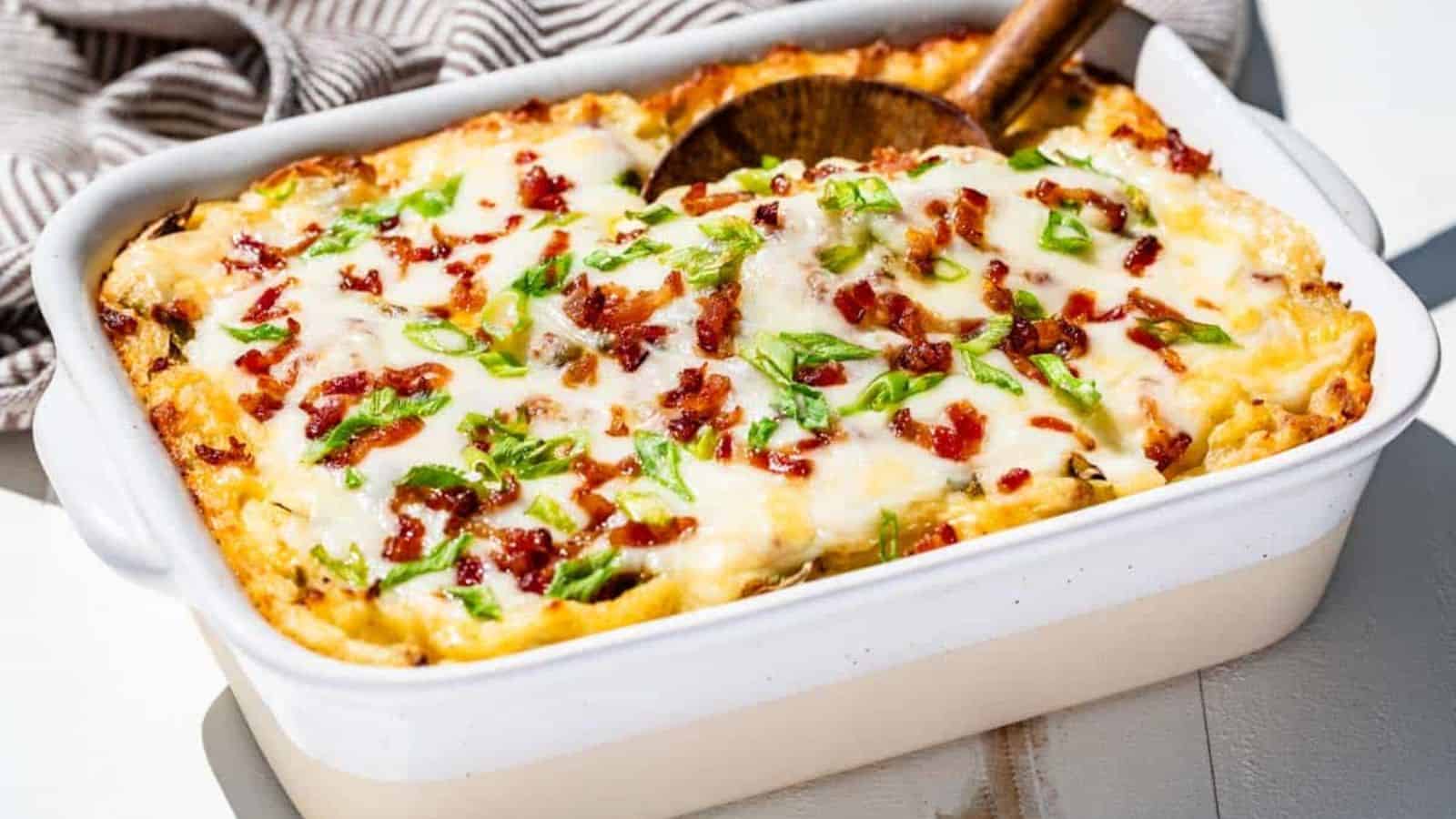 A baked casserole with melted cheese, crispy bacon bits, and green onion, served in a white dish on a neutral surface.