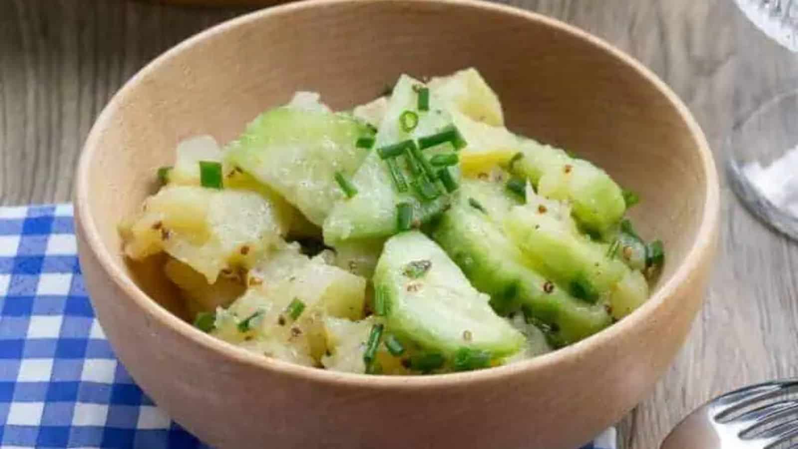 A wooden bowl filled with diced cucumber and potatoes, garnished with chopped chives, resting on a blue and white checkered cloth.
