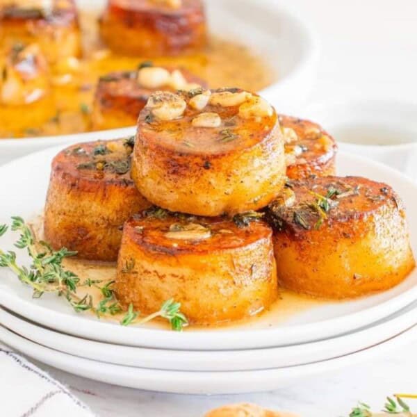 A stack of roasted potato rounds garnished with herbs sits on a white plate. Nearby, a sliced potato and forks are arranged, with a larger dish visible in the background.