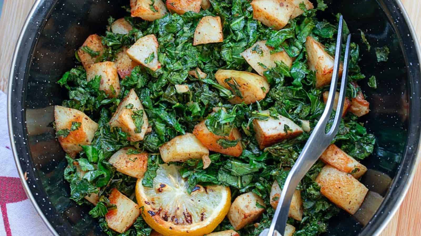 A bowl of sautéed potatoes and kale, garnished with a grilled lemon slice, featuring a fork resting on the edge.