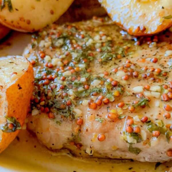 A close-up of a seasoned pork chop with mustard seeds and herbs, served alongside crispy potato wedges.