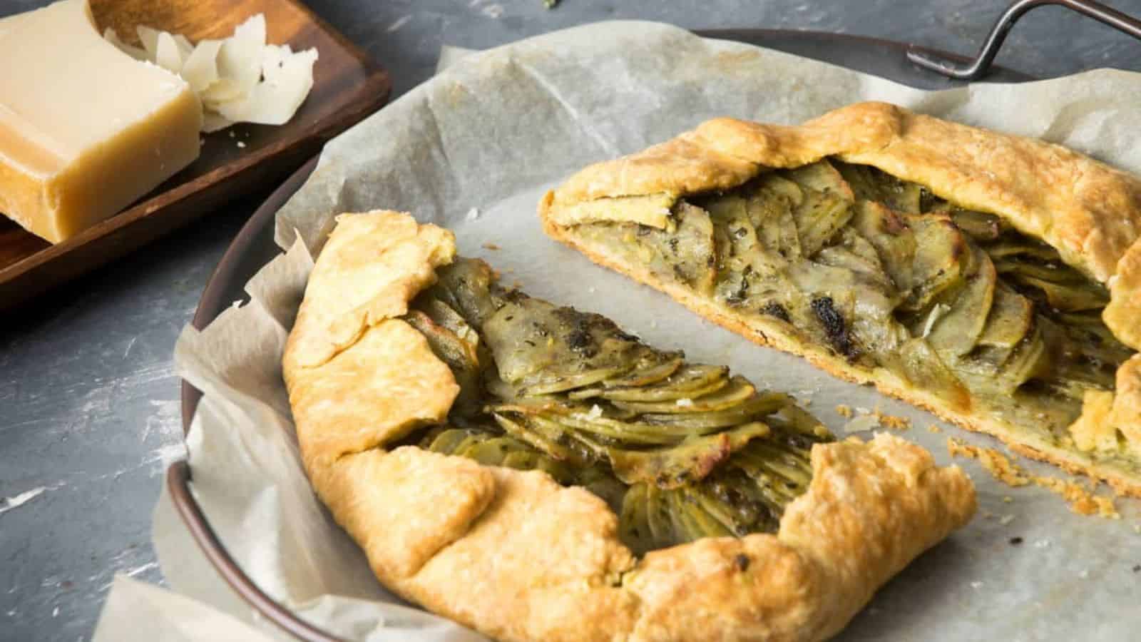 A golden-brown galette filled with layered vegetables displayed on parchment paper, alongside a wooden platter of cheese.
