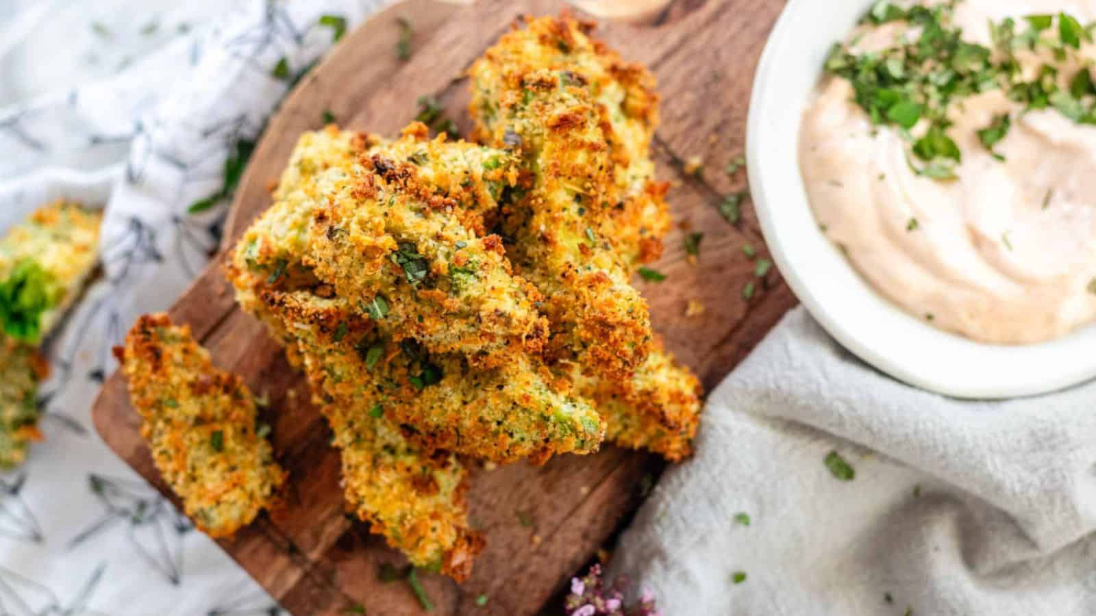 A wooden platter holding golden-brown vegetable sticks, topped with herbs, next to a bowl of creamy dipping sauce on a textured cloth.