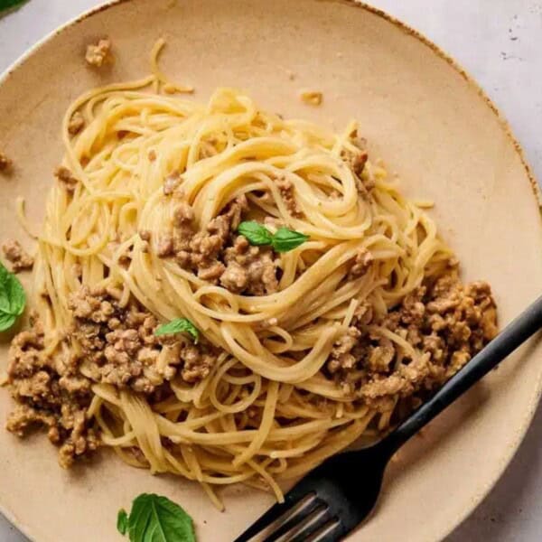 A plate of spaghetti with ground meat sauce, garnished with fresh basil leaves. A black fork rests on the plate's edge.