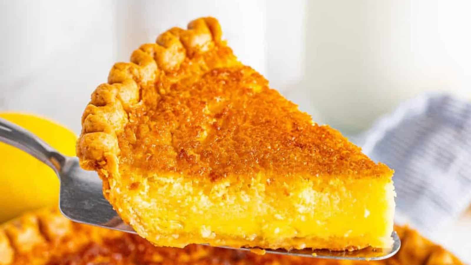 A golden slice of lemon pie with a flaky crust, held by a metal server, showcasing a creamy filling and a caramelized top.