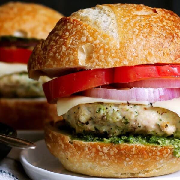 The image features a burger with chicken, cheese, tomato, onion, and green sauce on a bun, set on a white plate. Another burger is visible in the background.