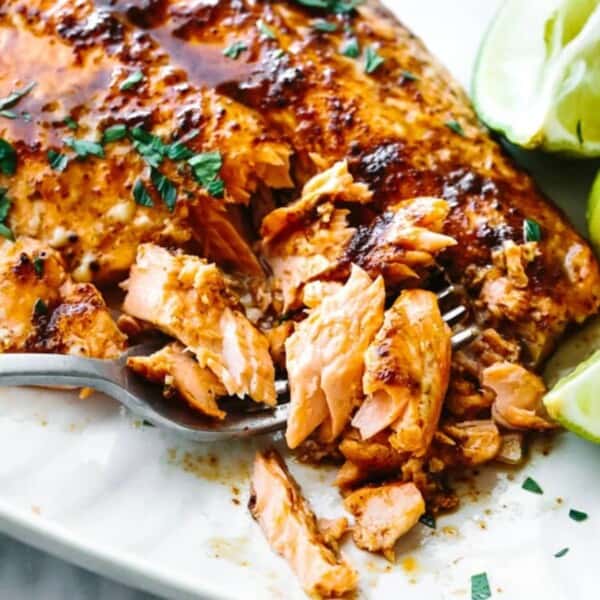 A grilled trout fillet garnished with herbs, lime wedges on the side, and a fork flaking the fish, all presented on a white plate.