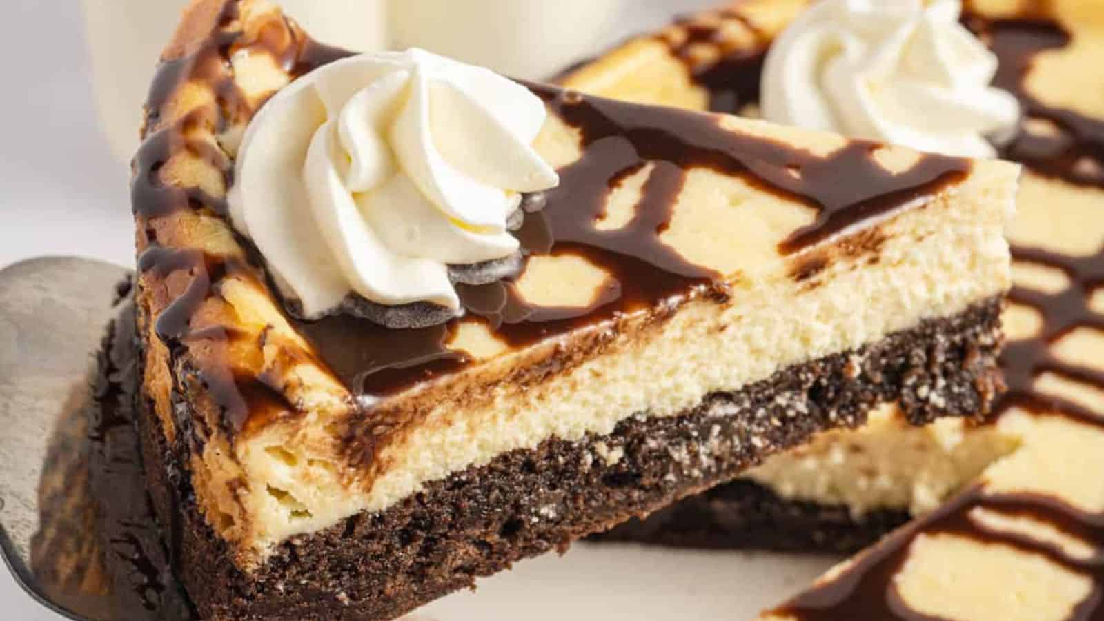 A close-up of a slice of cheesecake with a chocolate drizzle and whipped cream on top, sitting on a silver plate.
