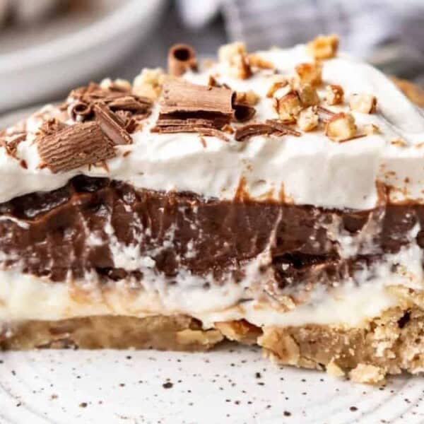A slice of layered chocolate and cream pie on a speckled plate, topped with whipped cream, chocolate shavings, and chopped nuts, with a fork on the side.