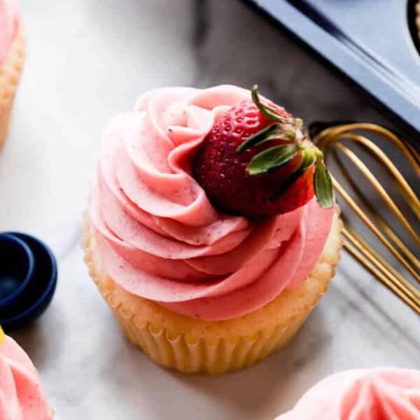 The image features a cupcake with pink frosting topped by a strawberry, along with a whisk and measuring spoons on a marble surface. Other cupcakes are nearby.