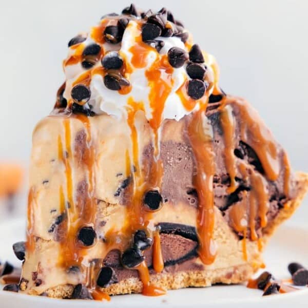 A slice of chocolate ice cream pie topped with whipped cream, chocolate chips, and caramel drizzle on a white plate.