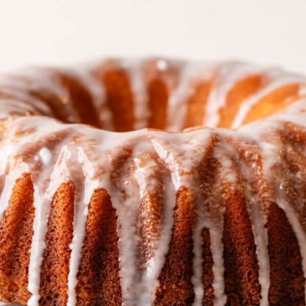 A bundt cake with a vanilla glaze drizzled over the top, forming a decorative pattern. The cake has a golden-brown crust and is placed on a white surface.