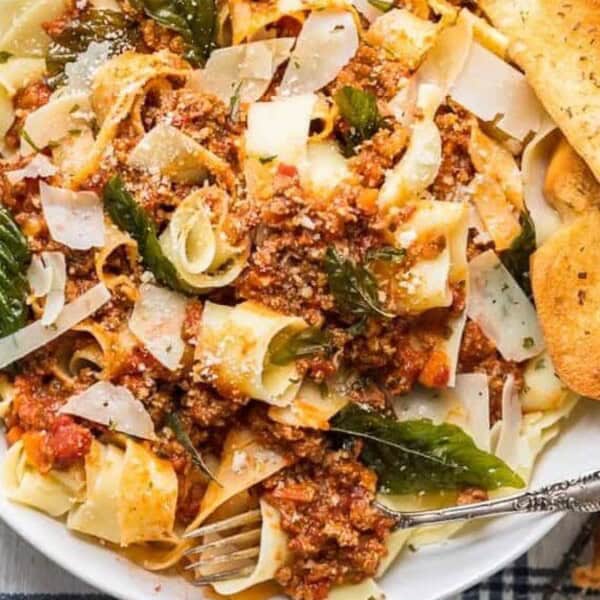 The image features a plate of pappardelle pasta topped with meat sauce, garnished with fresh basil and shaved cheese. Slices of garlic bread are placed nearby on a wooden table.