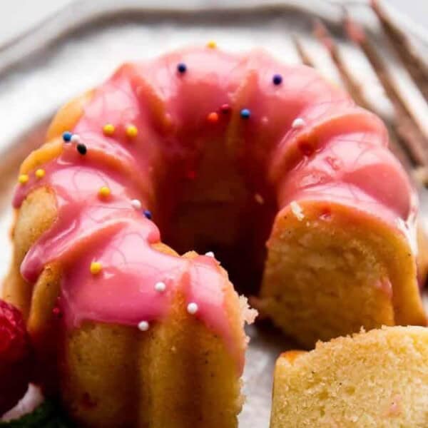 A mini bundt cake with pink icing and colorful sprinkles on a silver plate, accompanied by a fork and a raspberry.