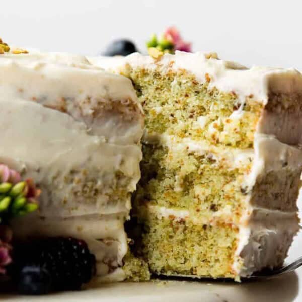 A three-layer cake with light frosting and a slice missing reveals a greenish interior. The cake is adorned with berries and small flowers on top and sides, set against a white background.