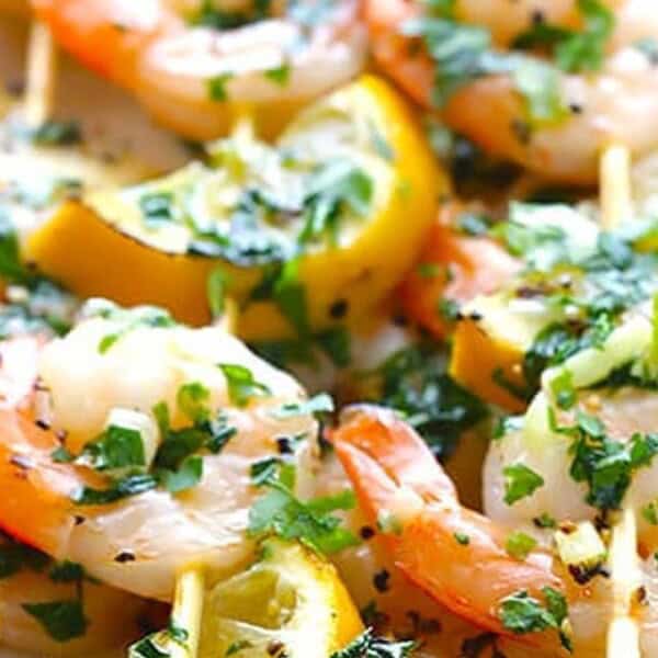 The image features grilled shrimp skewers accompanied by lemon slices, garnished with chopped herbs and sesame seeds, all arranged on a plate.