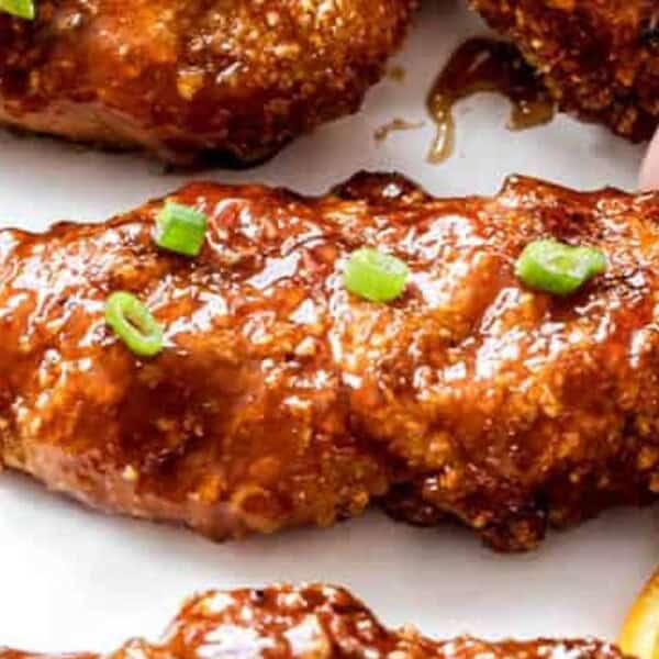 A glazed chicken tender with crispy edges is garnished with chopped green onions on a white plate. Juicy orange slices are placed nearby.