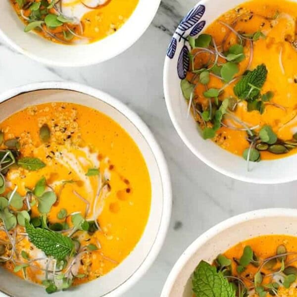 The image shows four bowls of vibrant orange soup adorned with greens and mint leaves. They are arranged on a table accompanied by spoons and fresh herbs.