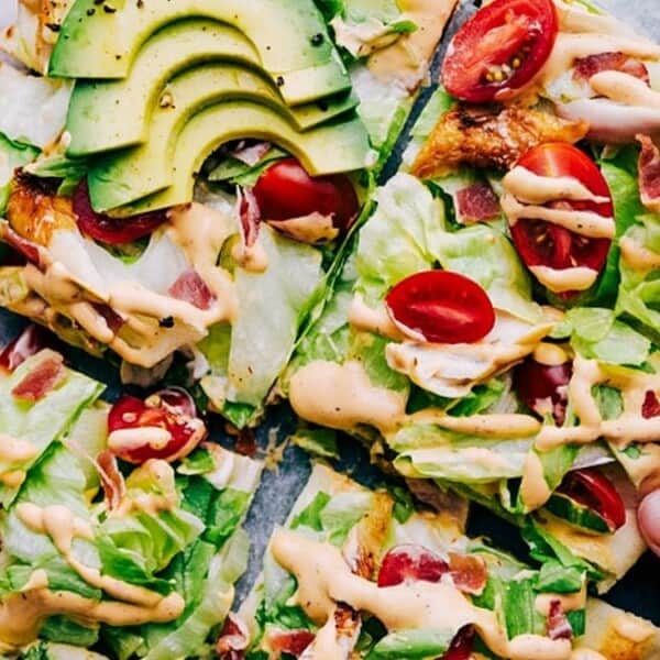 Pizza slices topped with avocado, cherry tomatoes, and lettuce, drizzled with sauce. A hand picks up a slice.