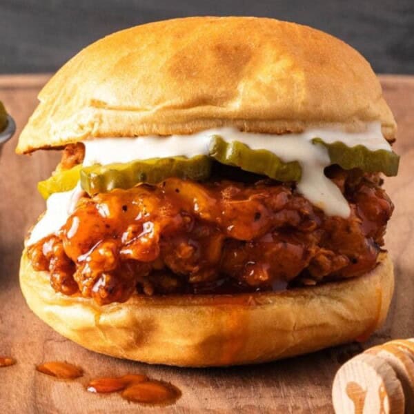 A chicken sandwich with pickles, white sauce, and a honey drizzle sits on a wooden board. A small cup of pickle slices is placed nearby.