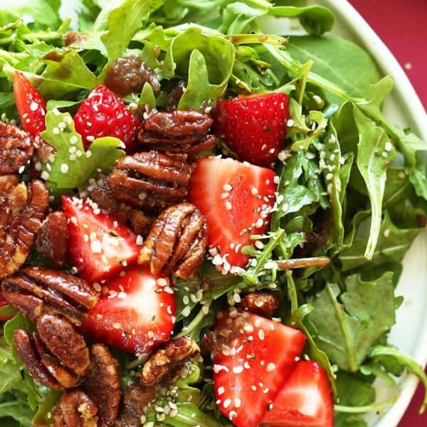 The image shows a salad composed of arugula, spinach, strawberries, pecans, and sesame seeds on a white plate. A fork and an additional strawberry are positioned beside the plate on a pink surface.