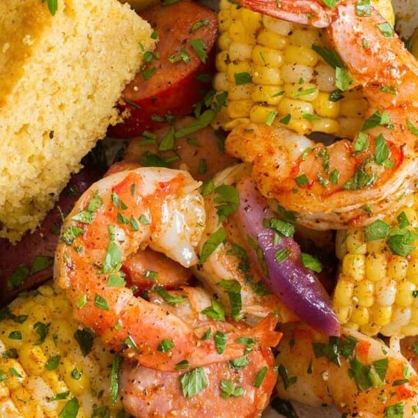 The image shows a plate containing shrimp, corn on the cob, sausage, potatoes, and red onion garnished with parsley. A piece of cornbread is served on the side.
